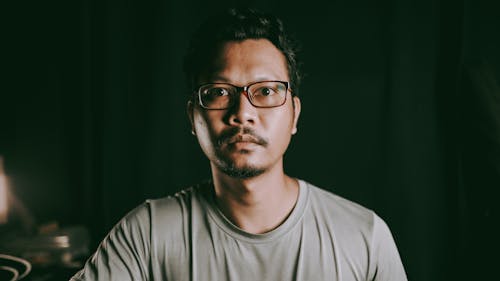 Emotionless bearded Asian male in casual shirt and eyeglasses sitting in dark room and looking away