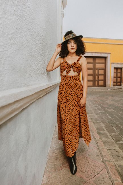 Fashionable woman with curly hair poses in an orange dress and hat against a textured wall.