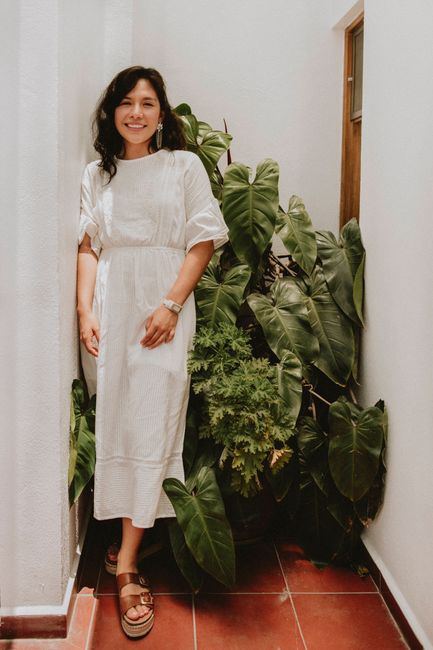 A smiling woman in a white dress stands indoors surrounded by lush green plants.
