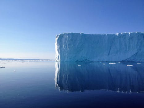 An awe-inspiring iceberg mirrored in calm Arctic waters under a clear blue sky.