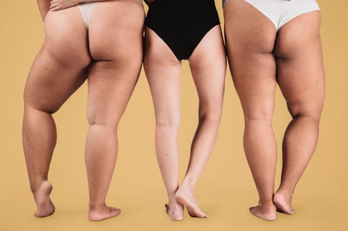 Back view crop anonymous females with slim and plus size figures wearing lingerie standing against beige background in studio