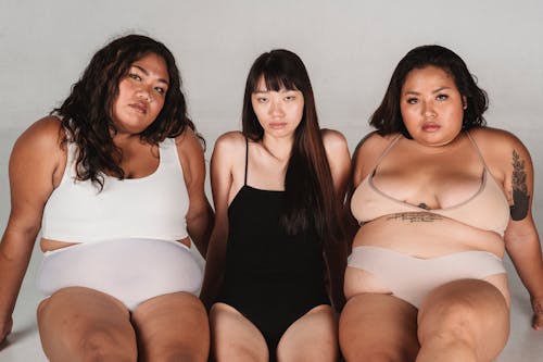 Calm Asian skinny woman wearing black bodysuit sitting on floor between plus size female models in light underwear against white wall and looking at camera