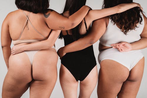 Back view crop anonymous females with slim and plus size body complexions in lingerie hugging each other shoulders and standing against white wall in photo studio