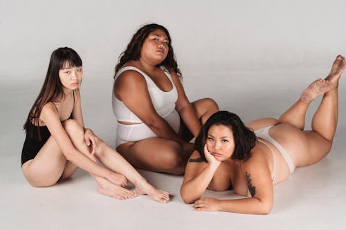 Full body emotionless Asian females with slim and overweight body complexions in lingerie resting on floor against white background during photo session in studio