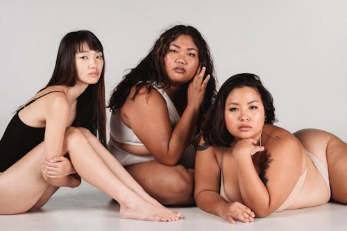 Feminine Asian females with fit and overweight body complexions wearing lingerie and sitting together on floor against white wall in light studio