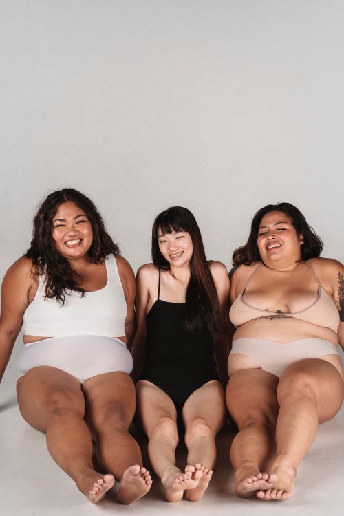 Free Smiling Asian females with different body complexions wearing lingerie sitting on floor and leaning on hands during photo session in light studio Stock Photo