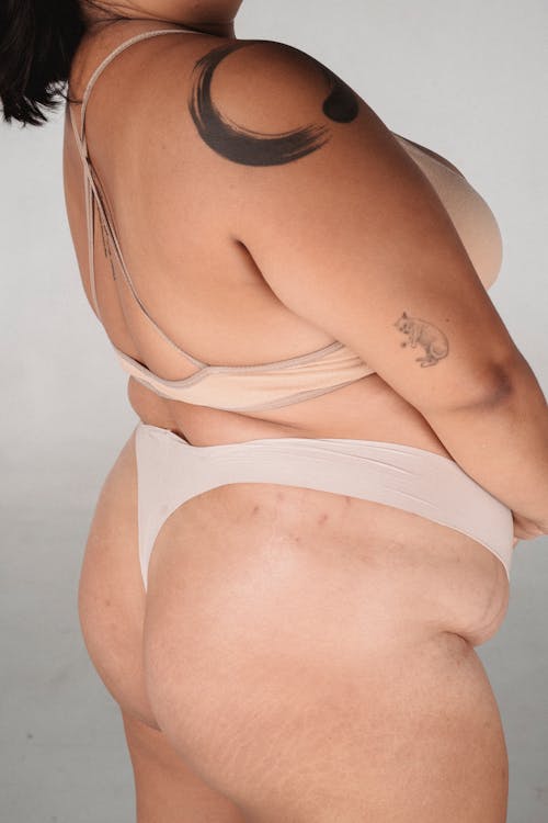 Crop plus size woman with tattoos in lingerie standing in studio on gray background