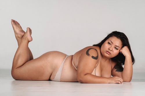 Full body of barefoot plus size Asian female with tattoo on shoulder in underwear lying on floor on stomach with feet in air on white background