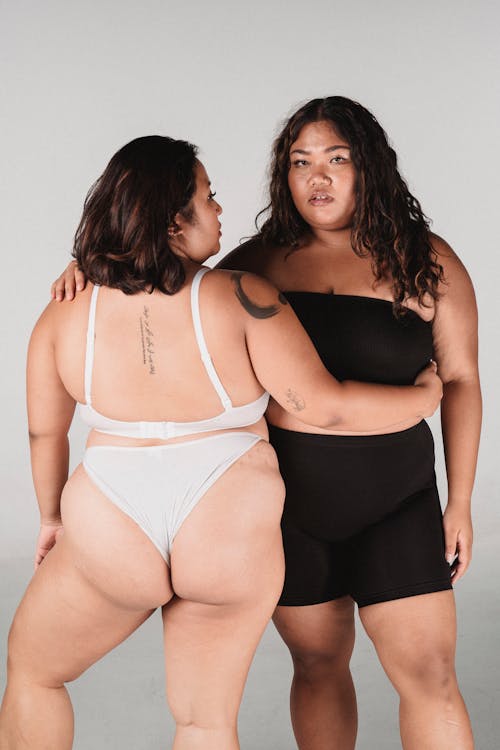 Back view of overweight woman wearing thong embracing plump Asian female while standing on white background and looking at camera