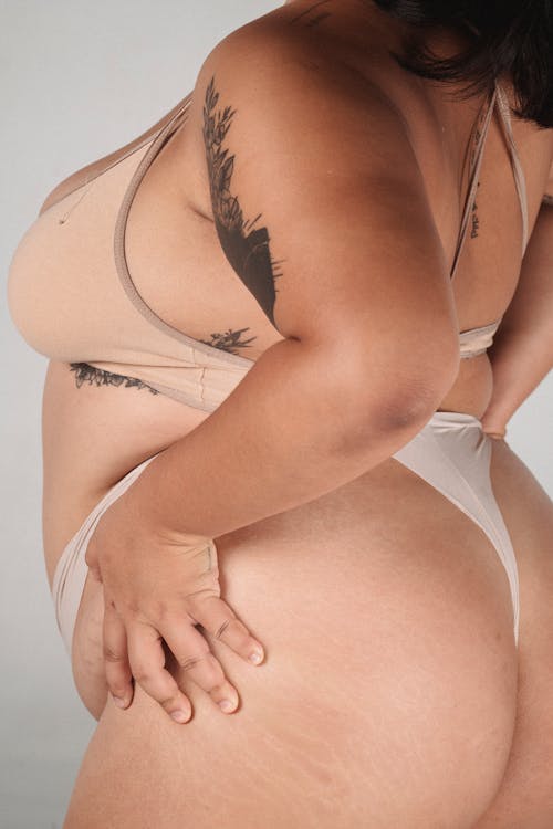 Side view of plus size female with tattoos wearing bra and thong standing with hands on hips on white background