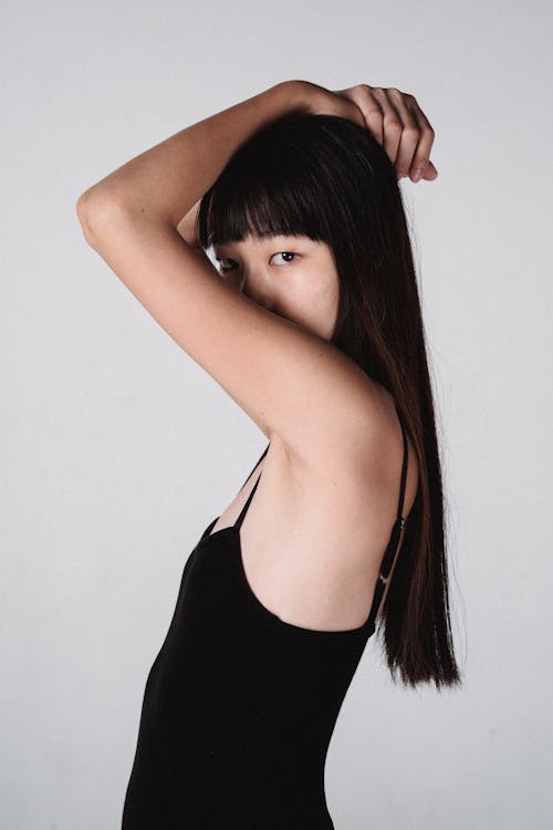 Slim ethnic female in black underwear standing in studio while covering half of face and looking at camera