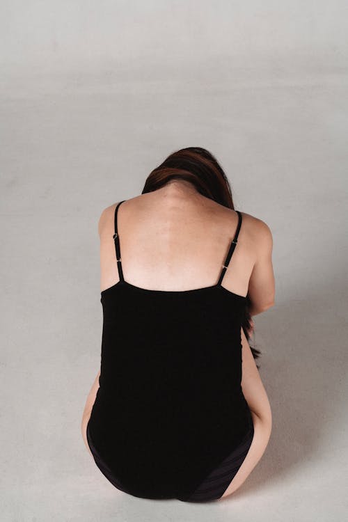 Back view of slender faceless female in black underwear sitting on floor in studio