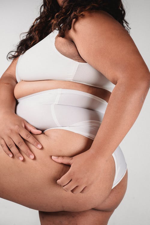 Unrecognizable plus size female in white underwear standing on white background in studio with raised leg and touching skin on hip