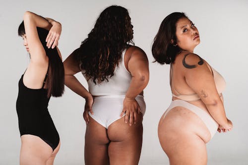Slim female with hands behind back standing in studio with plus size Asian models in white underwear on white background