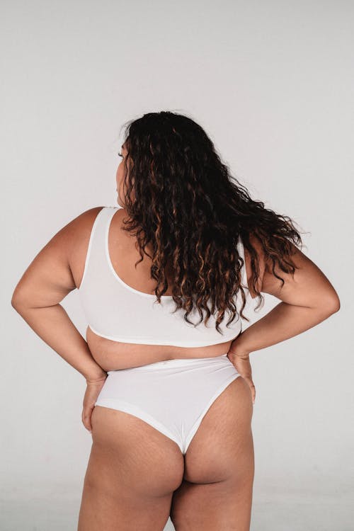 Back view of plus size anonymous female with curly hair wearing white lingerie while standing on white background with hands on waist