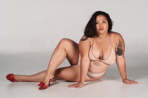Full body of plus size female with tattoos on body wearing shoes and lingerie looking away while sitting on white background with crossed leg