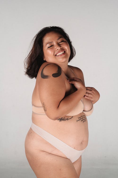 Plus size ethnic cheerful female with tattoo touching breast and smiling on white background