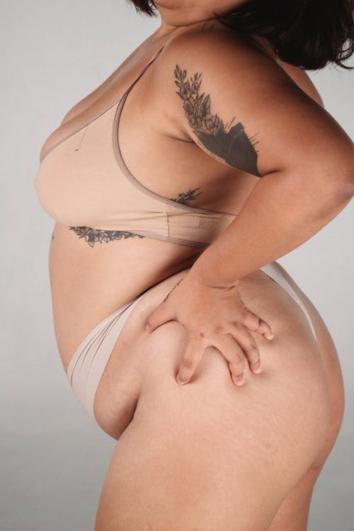 Side view of crop anonymous tanned plus size female with tattoo squeezing body on white background