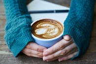 在舒适的环境中，木桌上放着一杯拿铁，从上方可以看到拉花艺术，旁边还有一只穿着毛衣的手