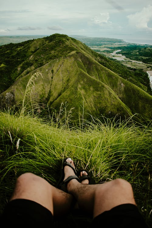 A Person Wearing Hiking Sandals Sitting on Grass Field in Front of Mountain View