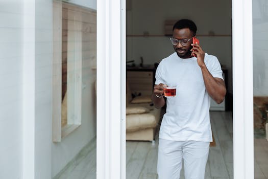 Stylish man indoors with eyeglasses, holding tea while talking on phone.