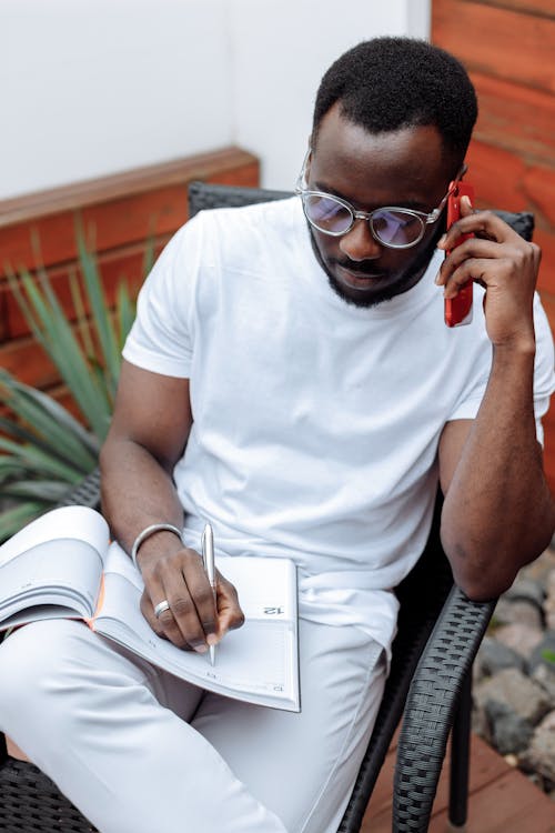 A Man Writing on a Planner While Holding His Smartphone 