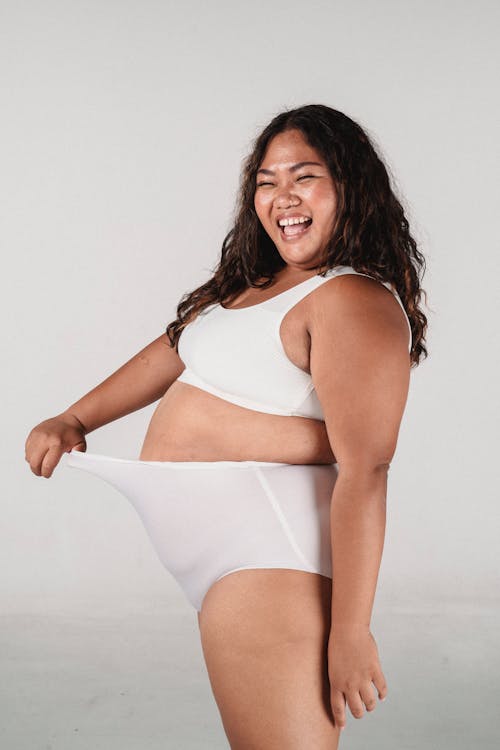 Side view of plus size female model in bra touching panties while laughing against white background
