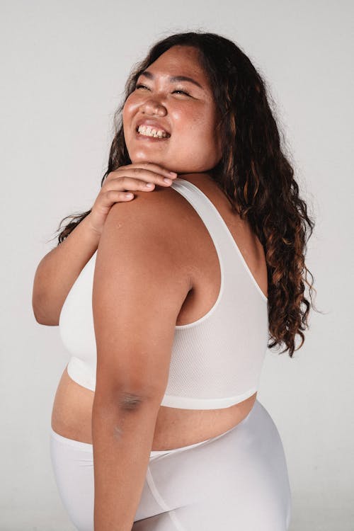 Side view of happy plus size female model in white bra and panties holding hand on shoulder and laughing while standing in studio