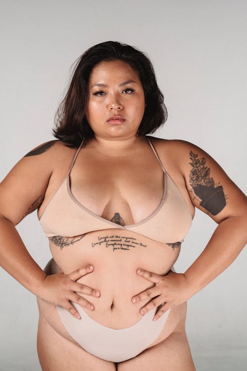Calm plus size Asian female showing curves of body while standing with hands on waist against gray background