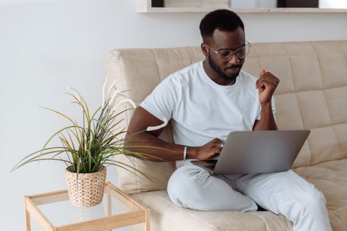 Free Confident young man sitting on a sofa using a laptop in a cozy home office environment. Stock Photo