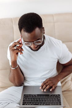 African man multitasking at home office, working on laptop while talking on phone.