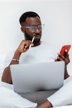Man thinking while using smartphone and laptop in home setting, eyeglasses visible.