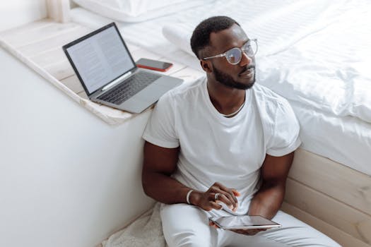 Man working from home with a laptop and tablet, reflecting in a bright room.