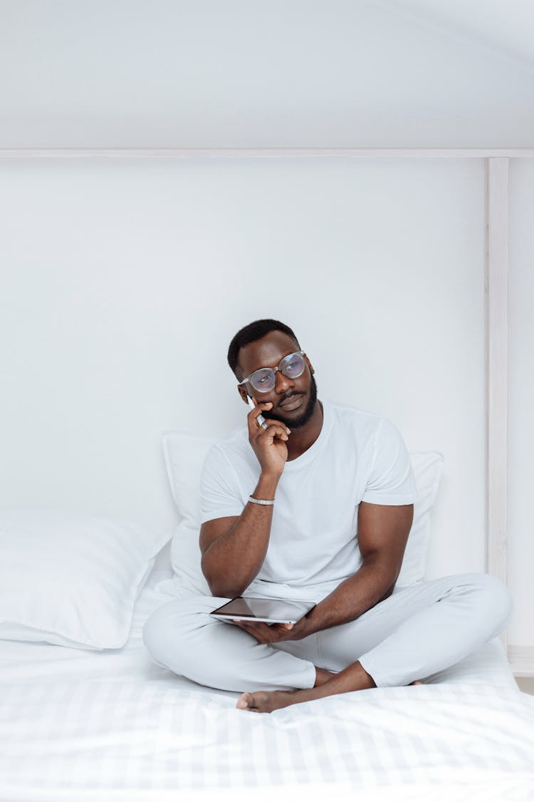 A Man Sitting On The Bed Thinking While Holding A Tablet And A Stylus Pen