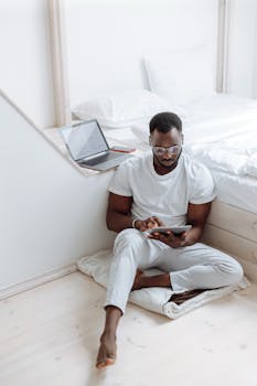 Man sitting on the floor, using a tablet for remote work effort next to a bed.