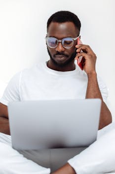 African American man wearing glasses, using phone and laptop in a bright indoor setting.