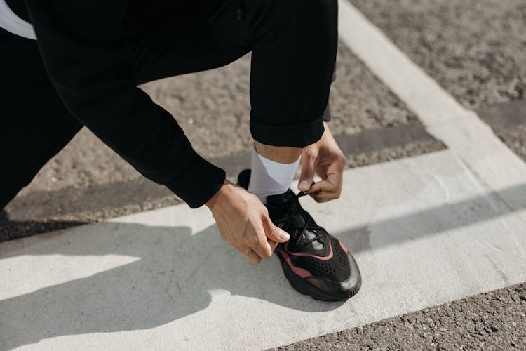 A Person Wearing Black Pants Tying The Lace Of The Sneaker He Is Wearing