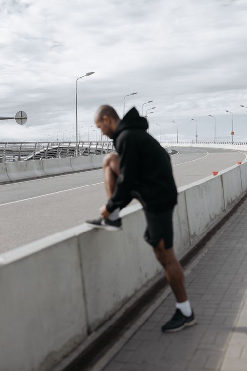 Free Man in hoodie tying shoelace on concrete barrier in urban environment, preparing for workout. Stock Photo