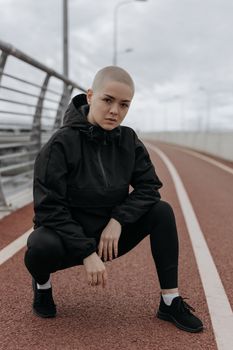 一名身穿黑色服装的秃顶女子蹲在桥上，展现了现代都市的时尚风格