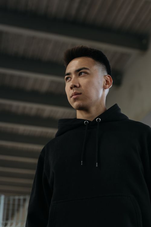 Free Portrait of a young Asian man in a black jacket, looking away under a modern urban structure. Stock Photo