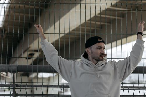 Free Young man in casual hoodie and cap by a chain link fence, outdoors under a bridge. Stock Photo