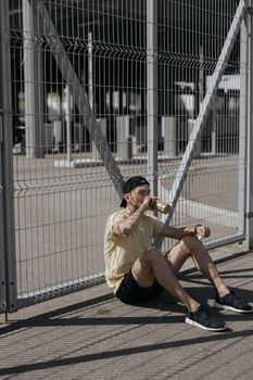 一个穿着黄色衬衫的年轻人在阳光明媚的户外，坐在栅栏旁喝酒