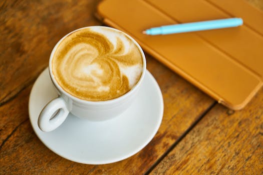 A white cappuccino cup with latte art sit on a rustic wooden table beside a notebook and pen.