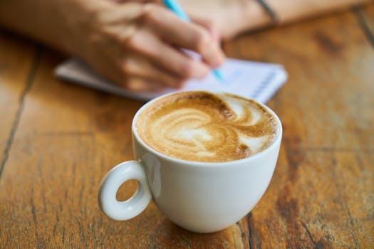 Close-up of a cappuccino cup on a wooden table with someone writing.