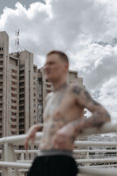 Shirtless tattooed man with urban high-rise backdrop and cloudy sky.