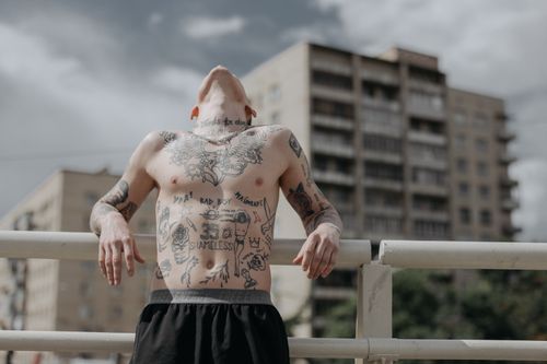 Free A shirtless man with tattoos leans against a railing with urban buildings in the background. Stock Photo