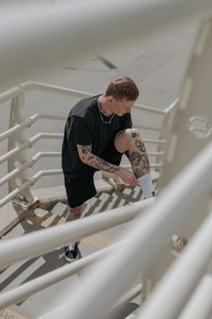Adult male with tattoos warming up on outdoor staircase, wearing sports attire.