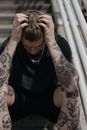 一名纹身男子穿着黑色衬衫坐在户外，双手抱头，看起来很疲惫