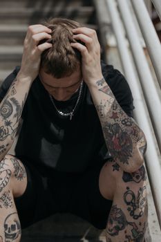 一名纹身男子穿着黑色衬衫坐在户外，双手抱头，看起来很疲惫