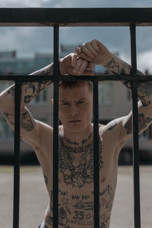 Free A tattooed man stands shirtless behind a metal fence in an outdoor setting, expressing a thoughtful mood. Stock Photo